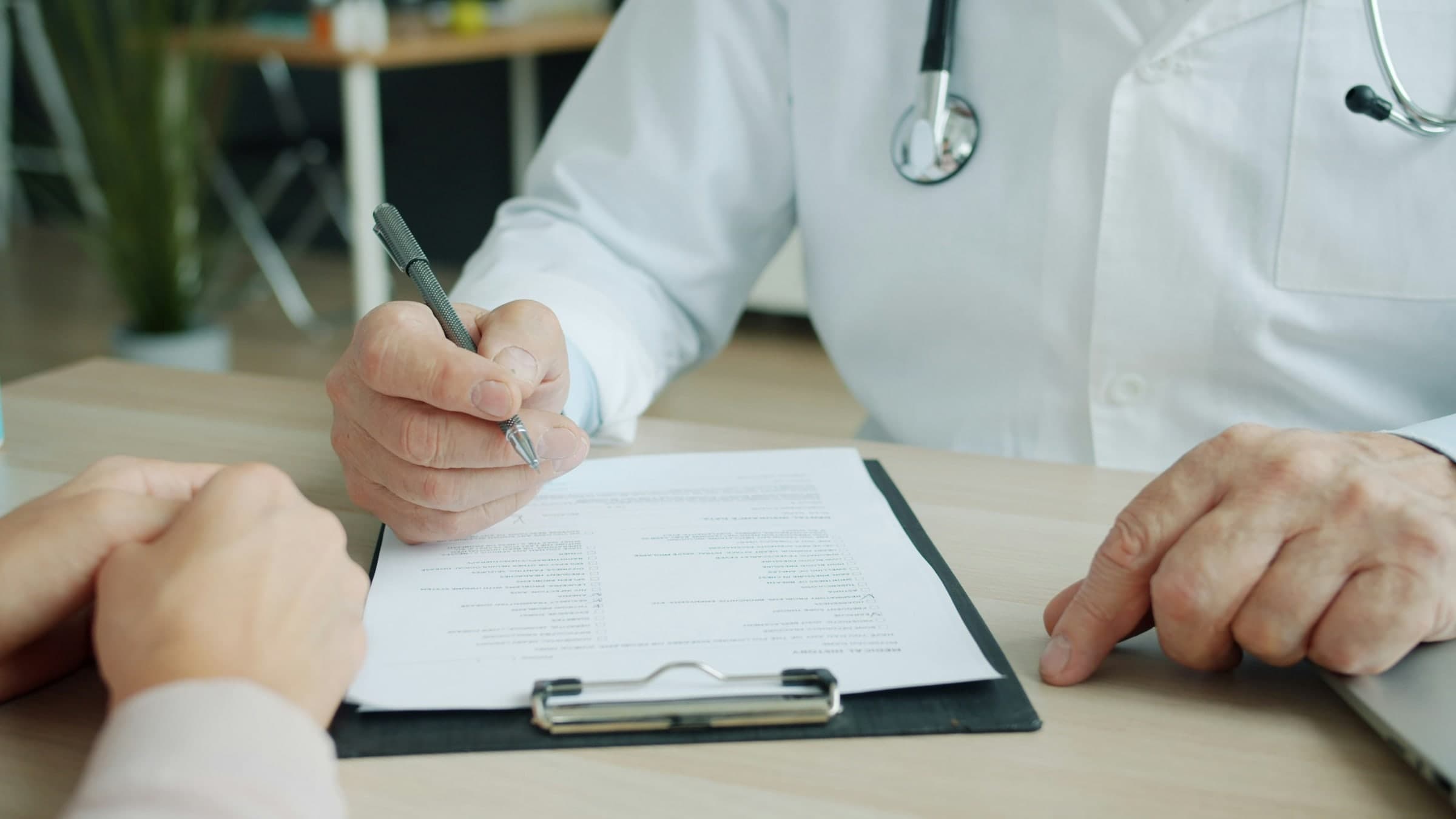 A clinician writes on a clipboard form while a patient sits across the desk.