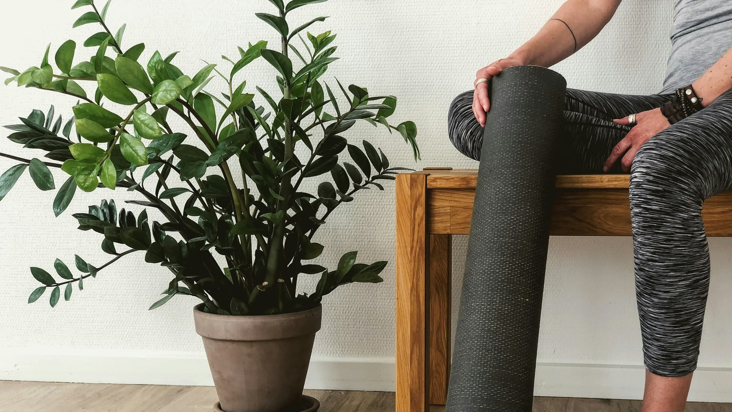 A person sits on a wooden bench at home, holding a rolled exercise mat beside a potted plant.