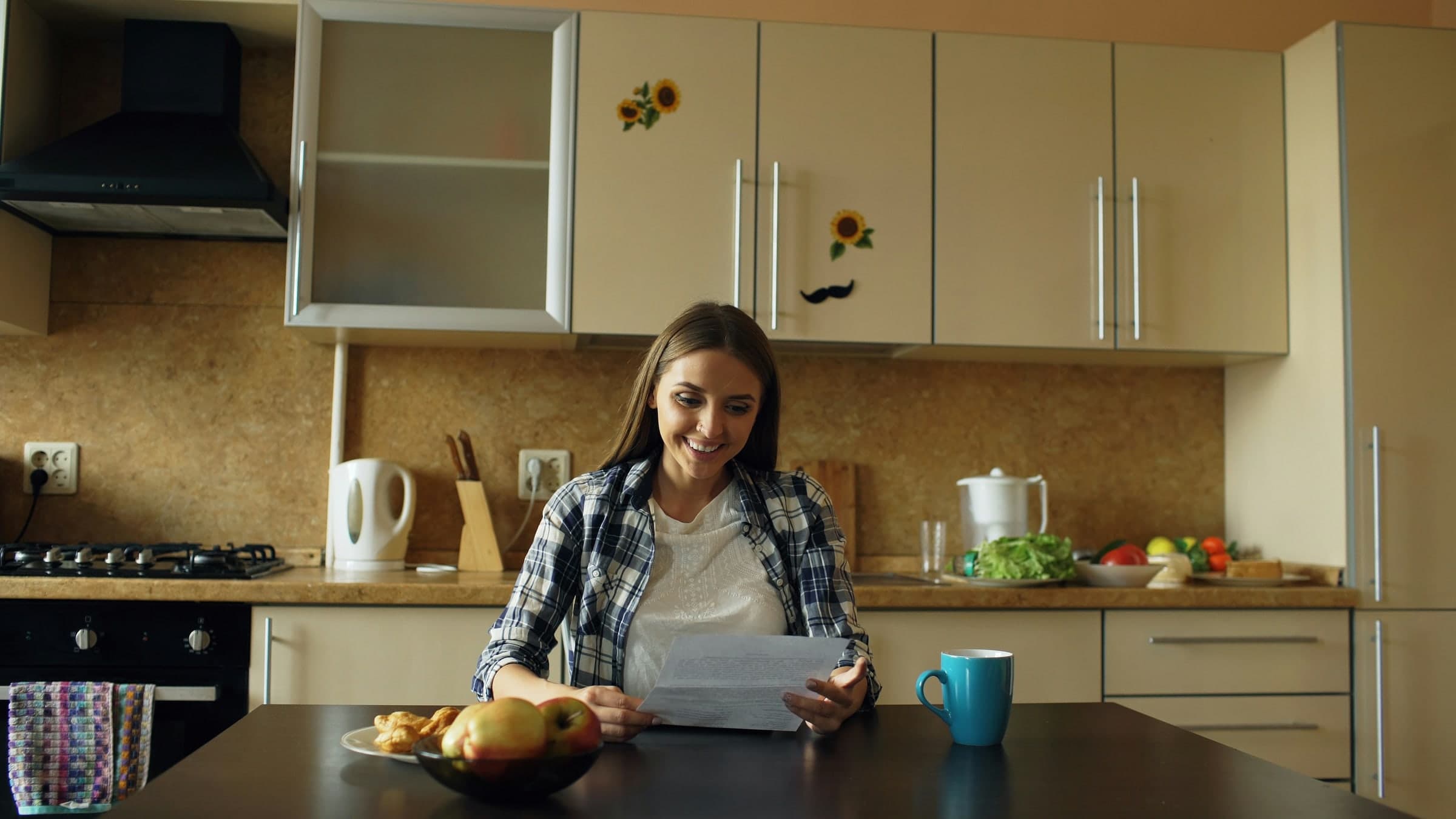 A smiling woman reads a single printed page at her kitchen table.